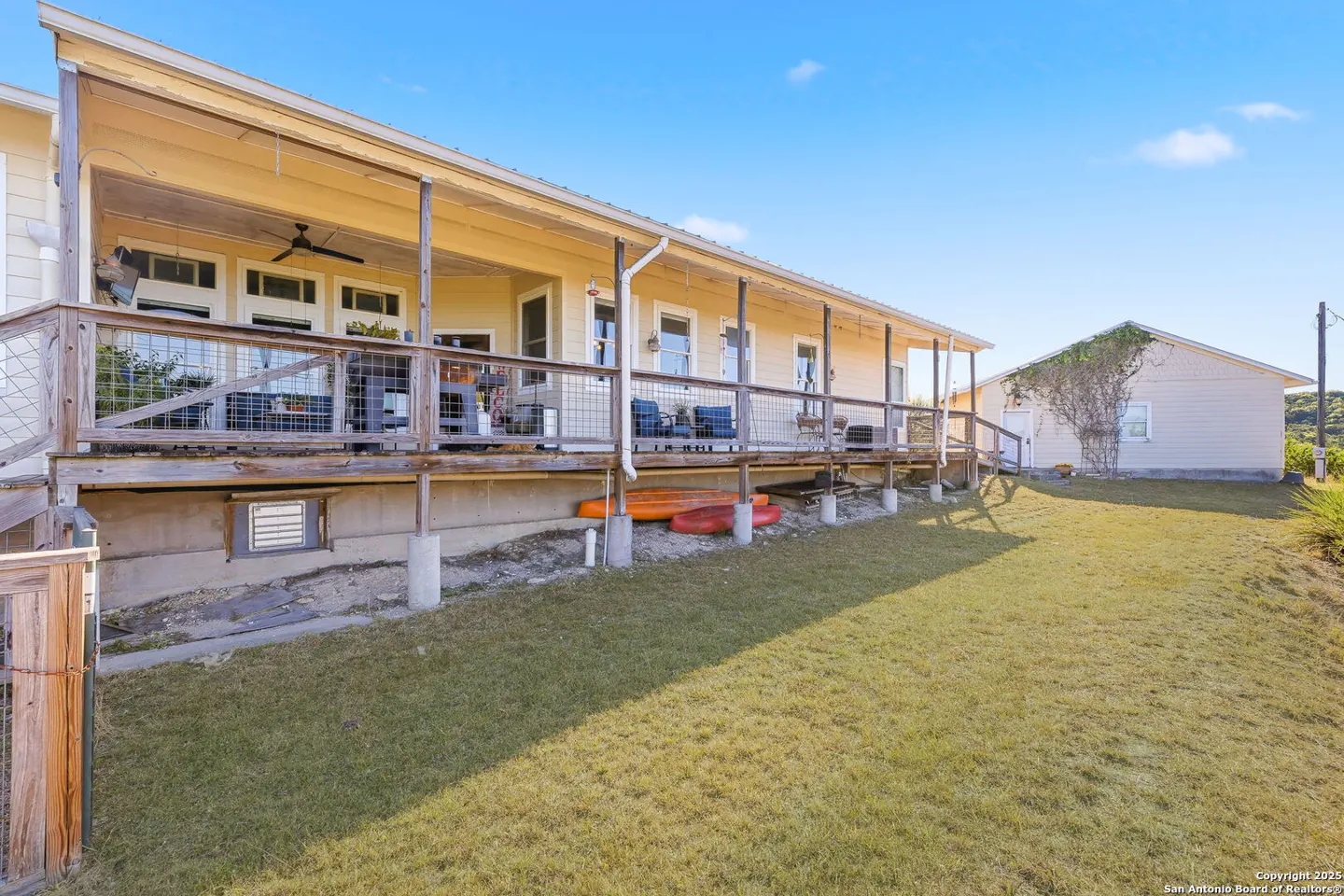 Tan house with a long porch, metal railing, and outdoor furniture. Orange kayaks are stored underneath. Green lawn and blue sky.