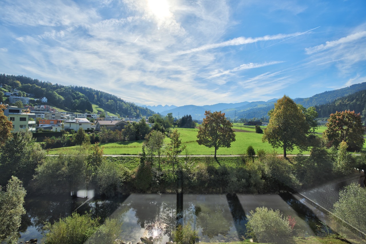 Scenic view of a river flowing past green fields, trees, and a small town under a bright blue sky with distant mountains. [AI-generated text]