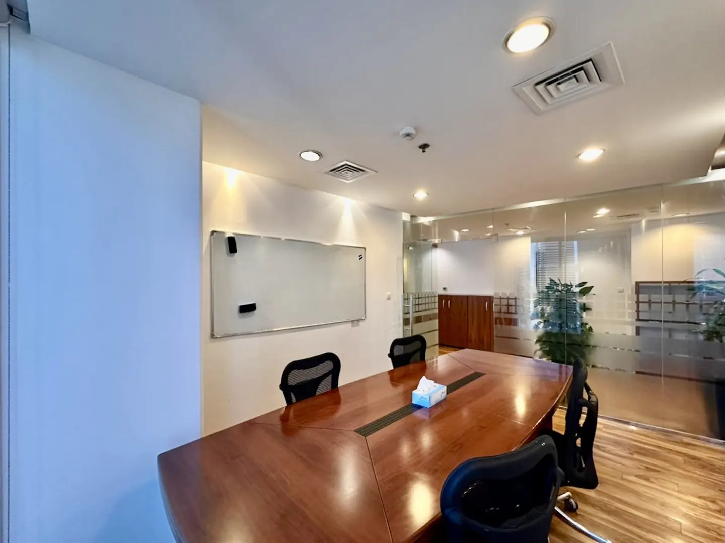 Modern office meeting room with a large wooden table, black ergonomic chairs, a whiteboard, and glass partition walls.