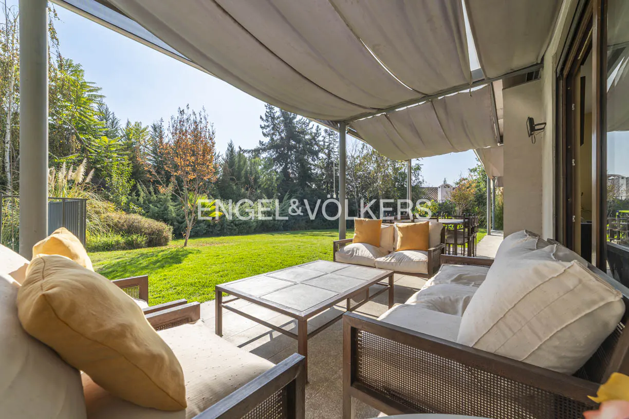Covered patio with outdoor seating, a table, and a view of a green lawn and trees. The Engel & Völkers logo is visible in the background.