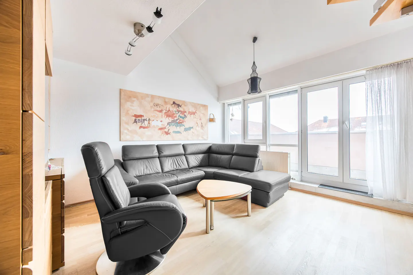 Bright living room with a black leather sectional sofa, chair, and a light wood floor. A large painting hangs above the sofa.