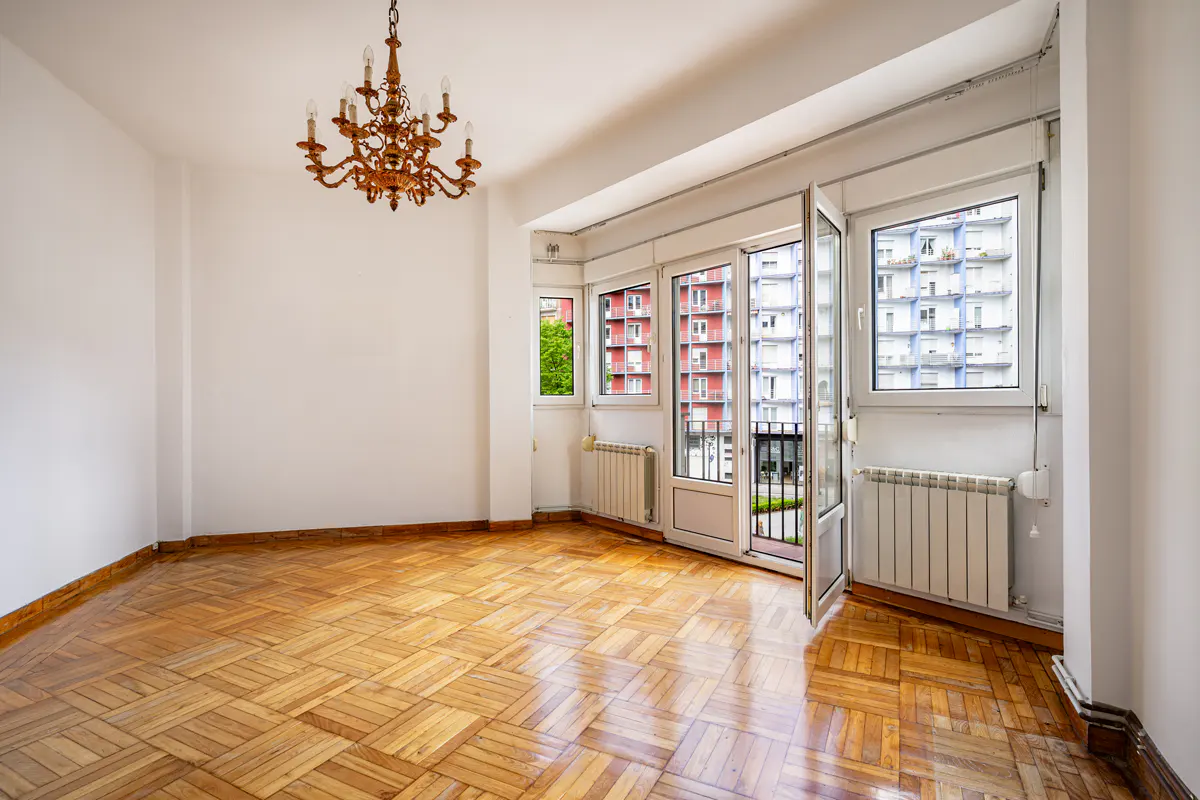 Empty room with parquet floor, white walls, and a chandelier. Balcony doors open to a view of buildings.