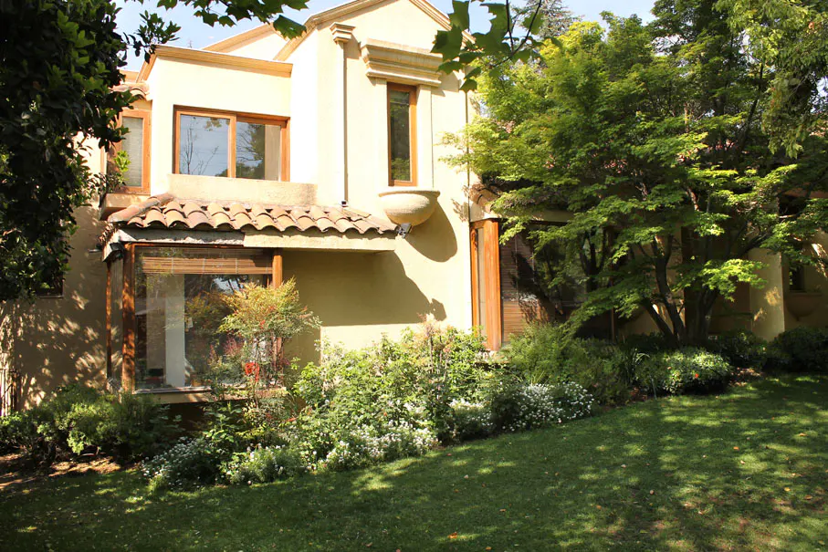 Two-story beige house with brown trim, red tile roof, and lush green landscaping.