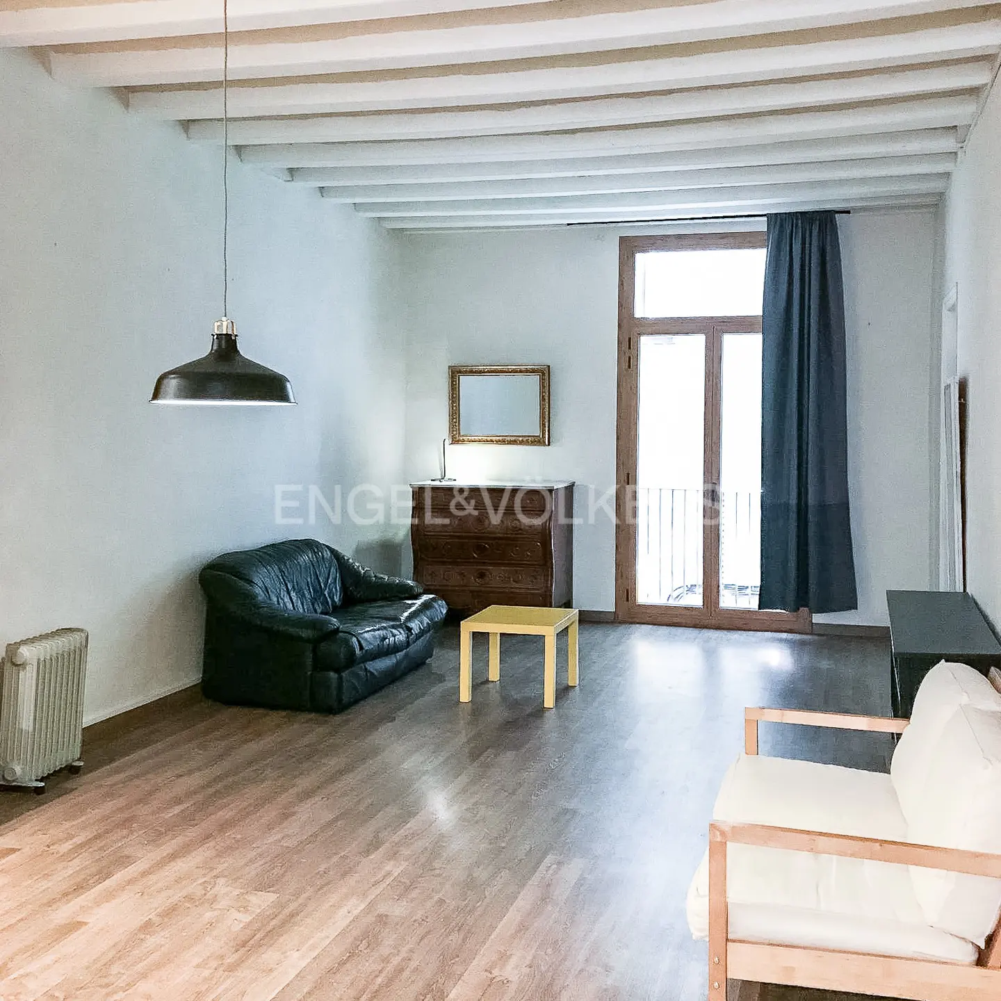 A spacious living room with a black leather sofa, wooden floors, and a white ceiling with exposed beams.