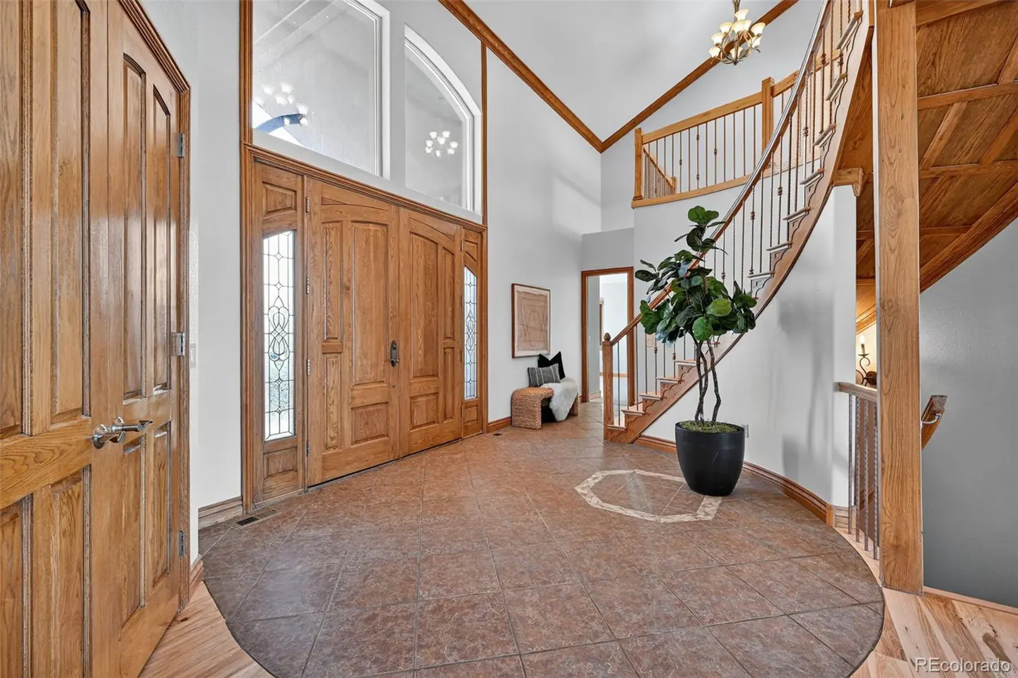 Grand foyer with wood doors, tile floor, and curved staircase. A large plant sits near the stairs. High ceilings and natural light.