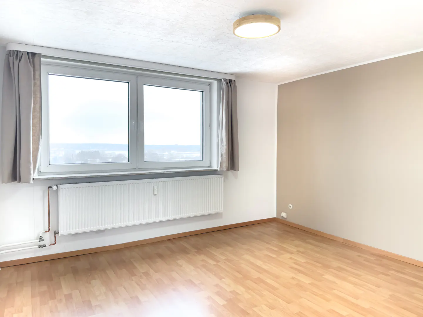 Bright, empty room with wood floors, a large window with gray curtains, and a white radiator. One wall is beige.
