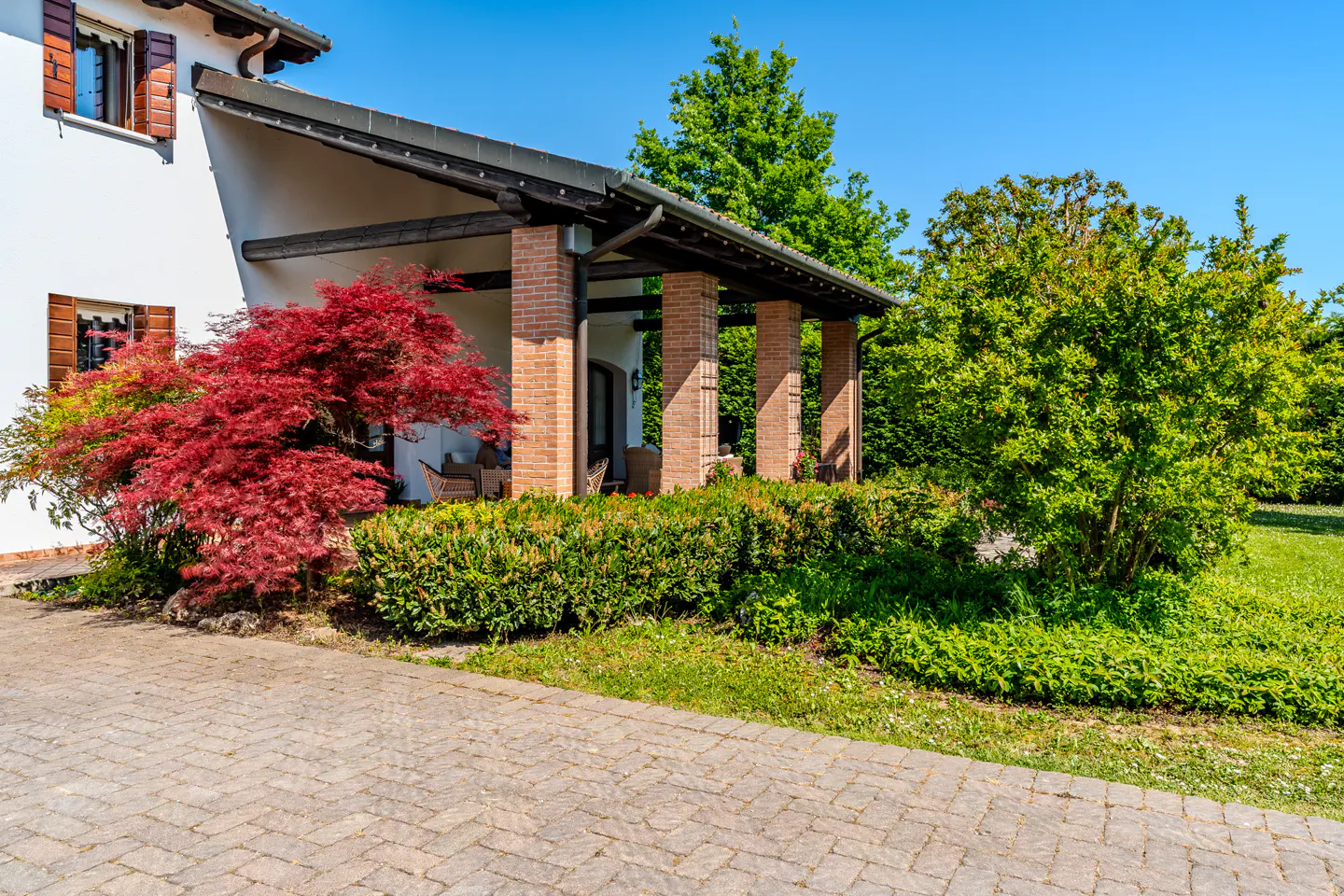 Exterior view of a white house with a brick columned porch, red Japanese maple, and green bushes under a clear blue sky.