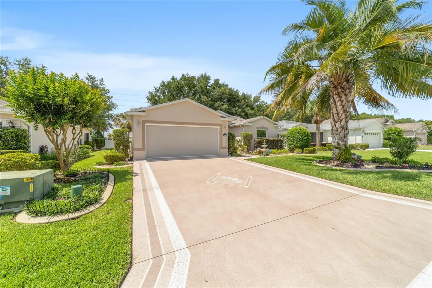 Beige single-story house with a two-car garage, long driveway, palm tree, and green lawn under a blue sky.