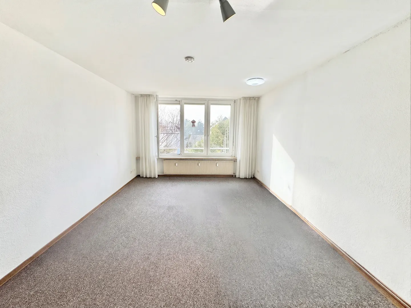 Empty room with gray carpet, white walls, and a window with white curtains. Two spotlights on the ceiling.