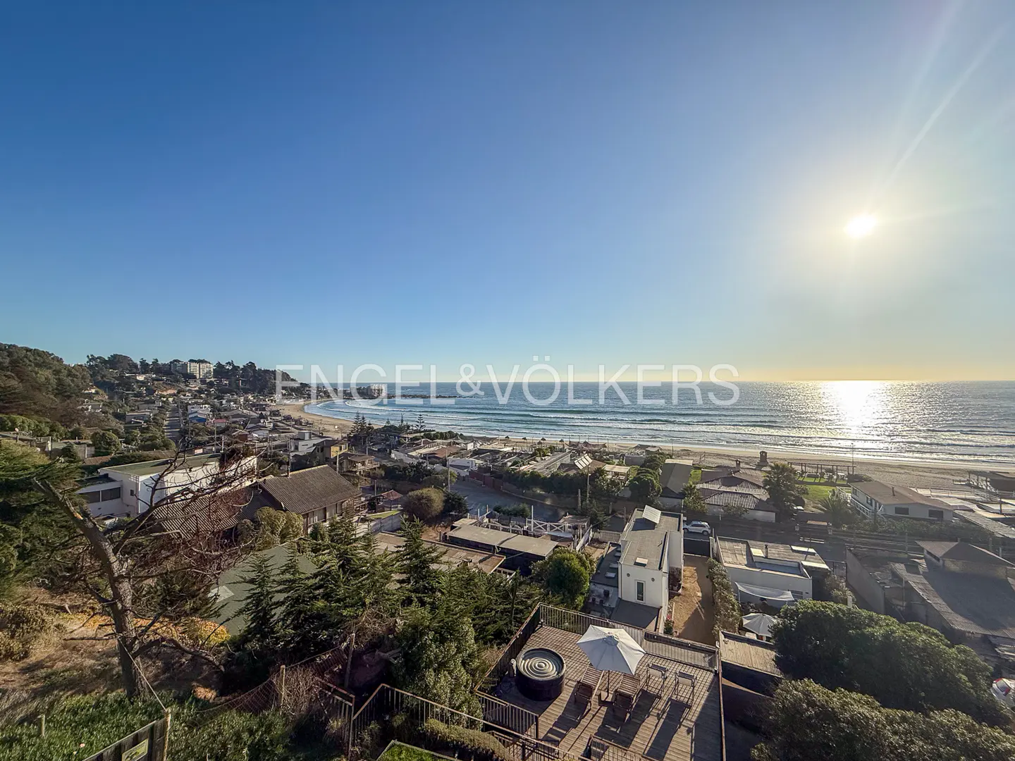 Ocean view from a deck with a hot tub, chairs, and umbrella. The beach and town are in the background under a blue sky.
