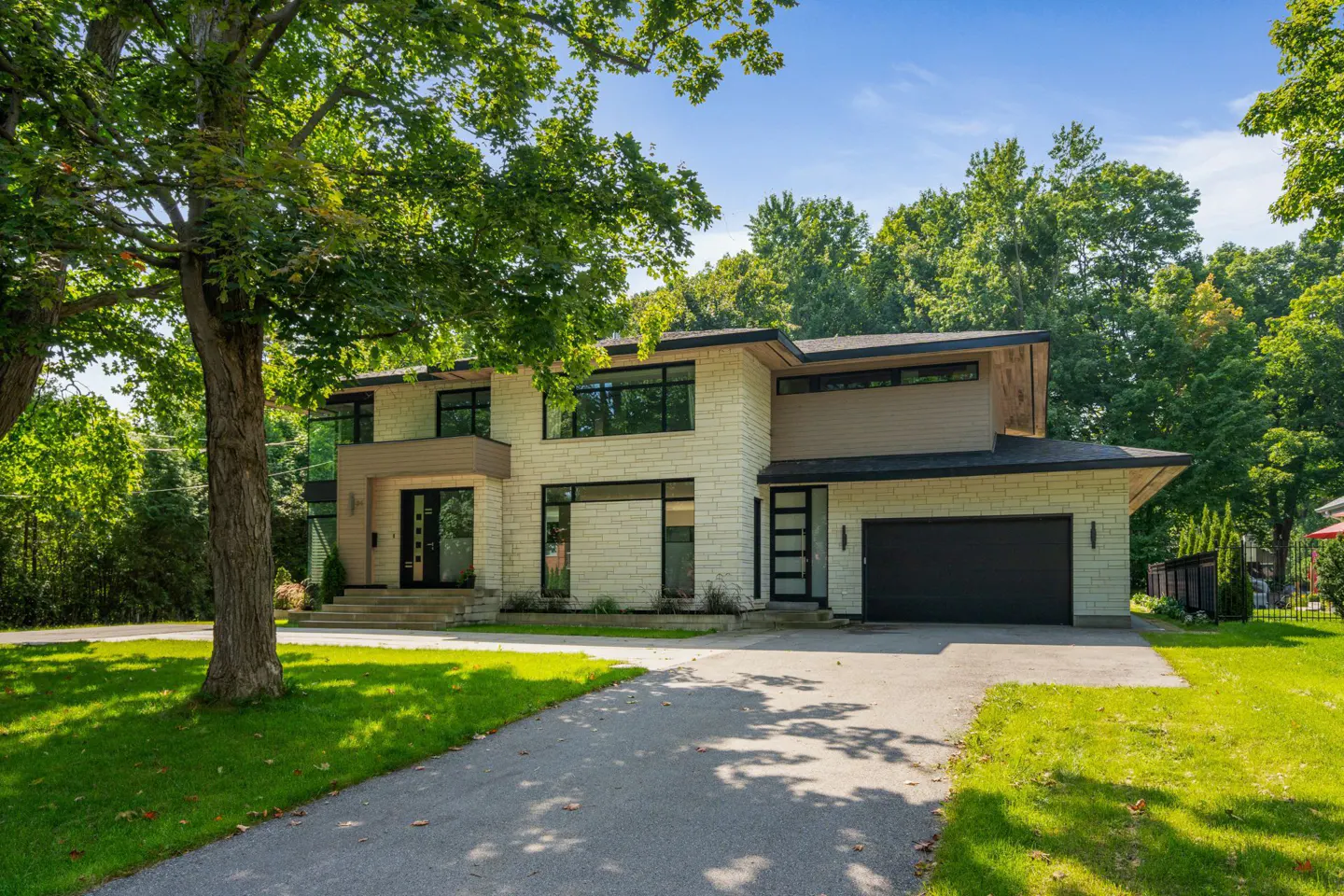 Two-story modern house with a black garage door, stone facade, and a long driveway. Green trees surround the house.