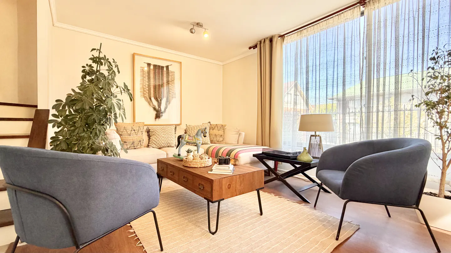 Living room with blue chairs, a wooden coffee table, and a white sofa with decorative pillows. Large windows with sheer curtains let in natural light.