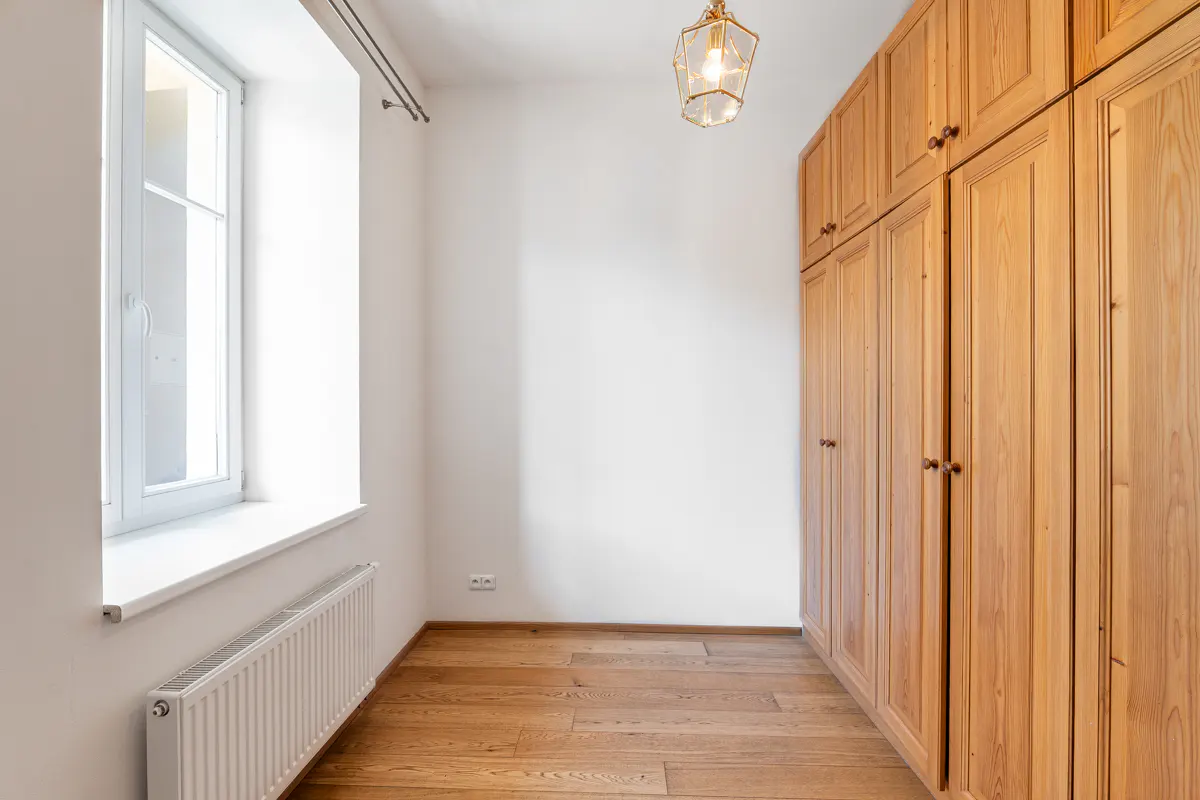 Bright room with wood floors, white walls, and a large window. A tall, light-wood wardrobe stands against one wall. A modern light fixture hangs from the ceiling.