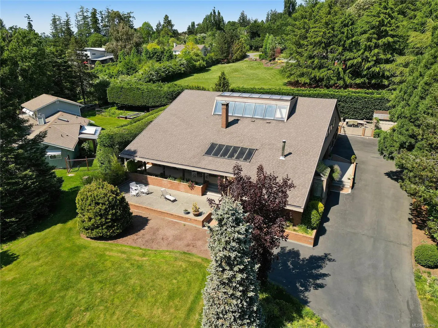 Aerial view of a single-story house with a brown roof, skylights, and a brick patio with outdoor furniture, surrounded by green lawns and trees.