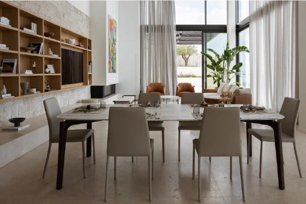 Bright dining room with a long marble table, gray chairs, and a built-in bookshelf. Large windows with sheer curtains let in natural light.