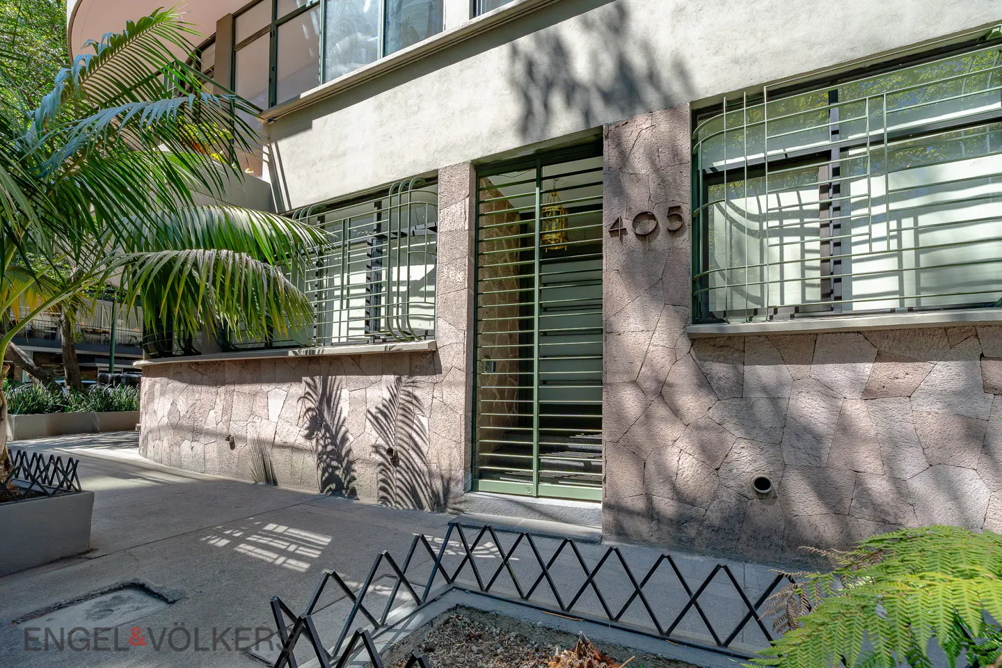 Exterior view of a building with the number 405 above a green door. Windows with metal bars, stone facade, and palm tree shadows.