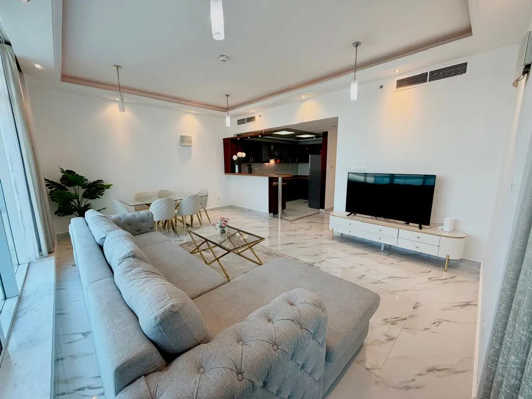 Modern open-plan living room with a grey tufted sectional, marble floors, dining area, and a kitchen in the background.