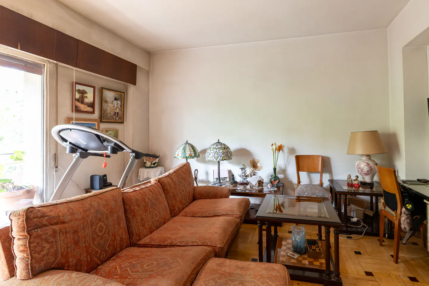Living room with a brown patterned sofa, treadmill, lamps, and tables on a parquet floor.