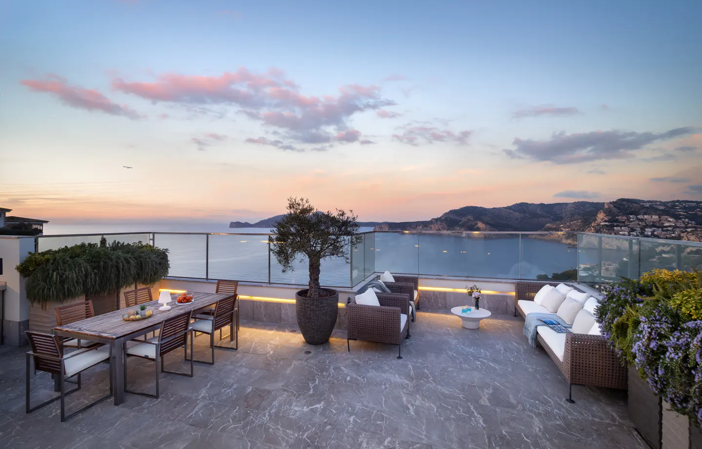 Luxury rooftop terrace with dining table, wicker seating, and ocean view at sunset.