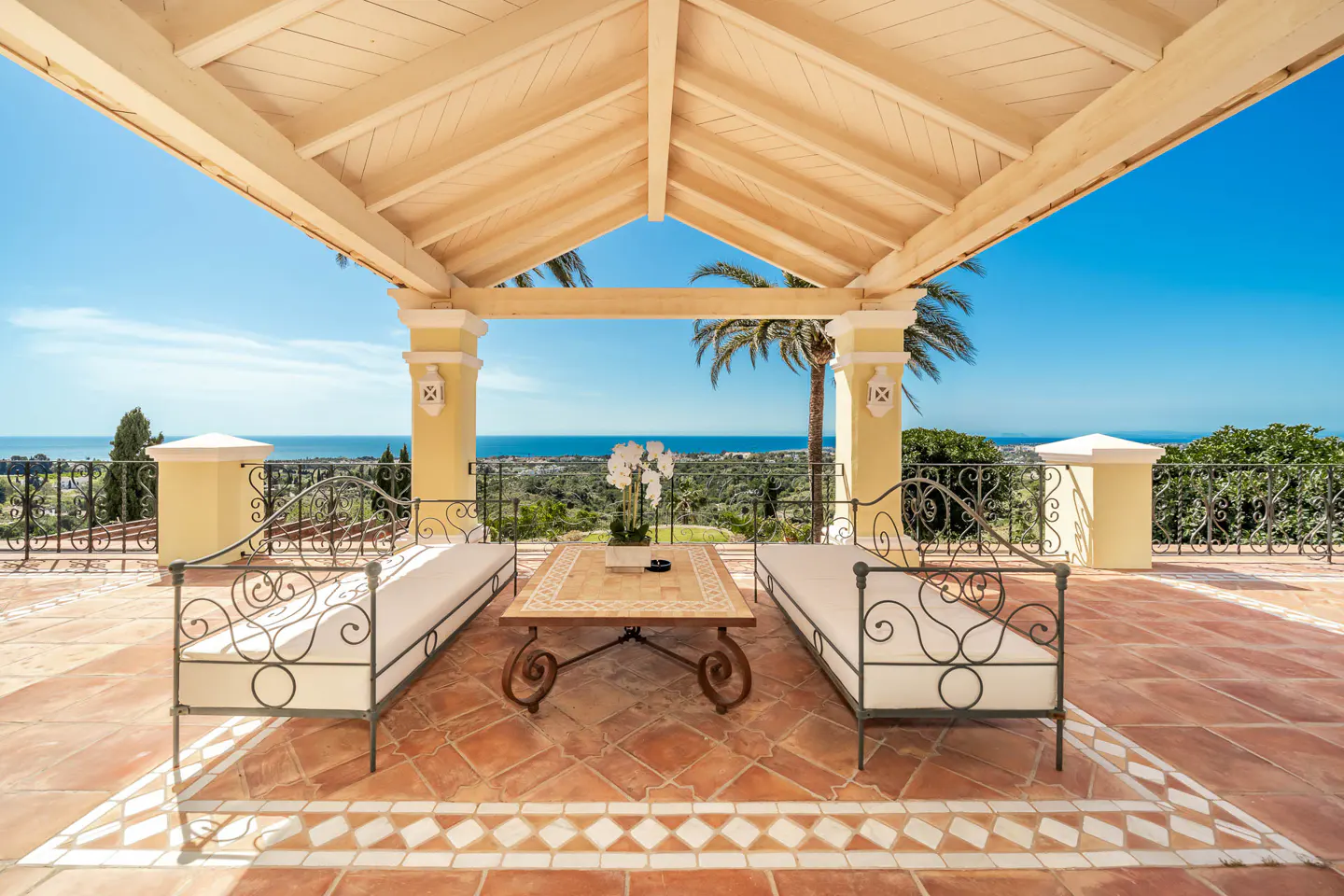 Outdoor patio with two white daybeds, a tile table, and a view of the ocean under a clear blue sky.