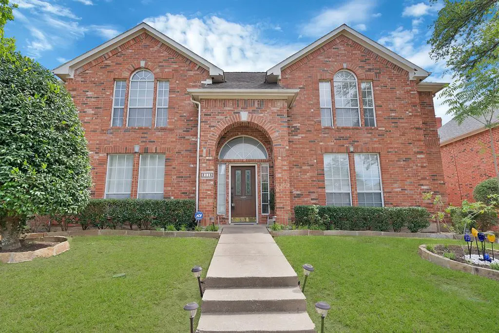 Two-story brick house with an arched entryway, a concrete walkway, and a green lawn under a blue sky.