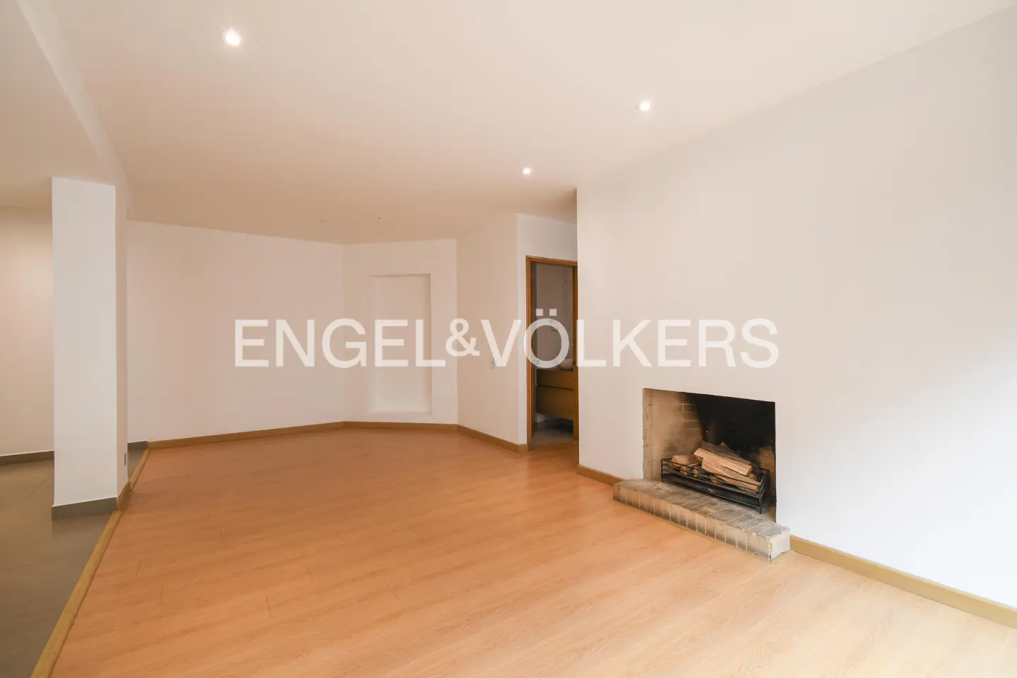 Bright, empty living room with wood floors, white walls, and a fireplace. Engel & Völkers logo visible.