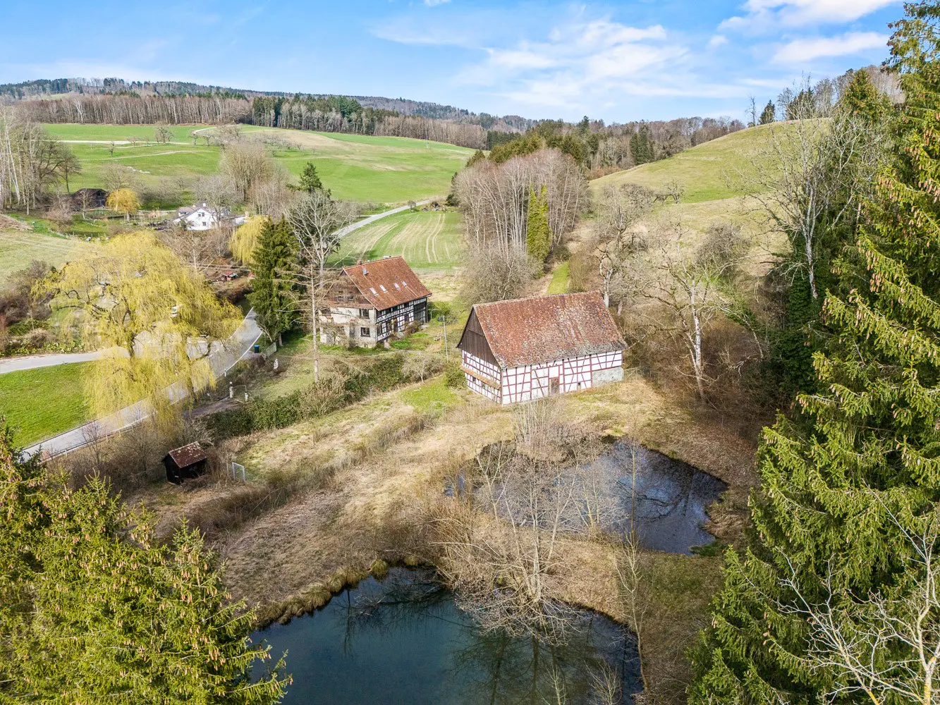 Aerial view of two traditional timber-framed houses with red roofs near small ponds in a lush, hilly rural landscape.