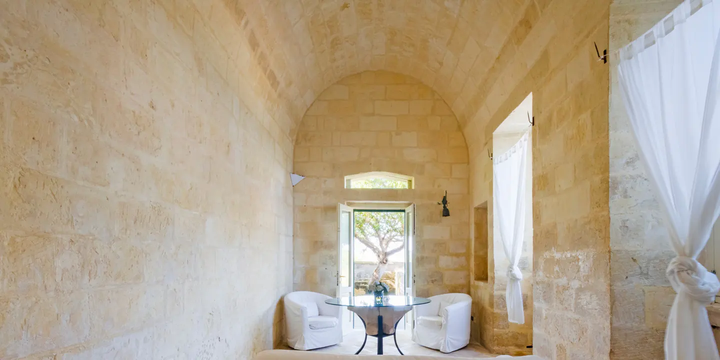 Interior view of a stone room with arched ceiling, white curtains, and a glass table with two white chairs. A tree is visible through the open door.