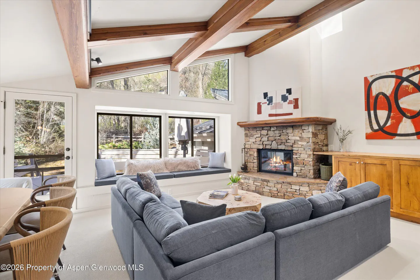 Living room with a gray sectional sofa, stone fireplace, and wooden beams on the ceiling. Large windows offer a view of the outdoors.