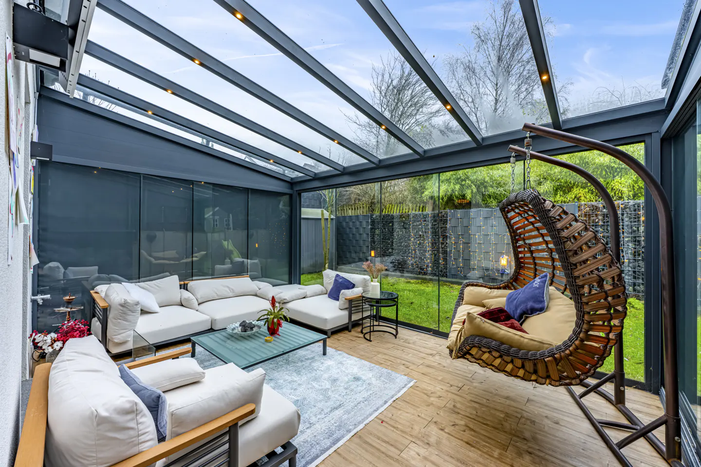 Sunroom with glass ceiling, white sofas, blue pillows, and a hanging wicker chair. Outside is a green lawn.