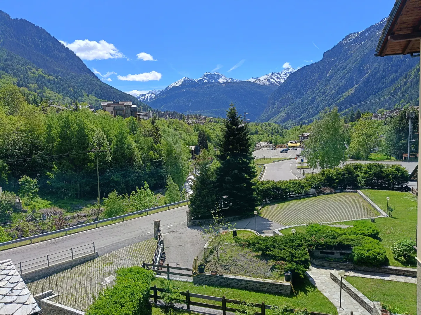 View from a balcony overlooking a valley with green trees, a road, and snow-capped mountains in the distance under a blue sky.