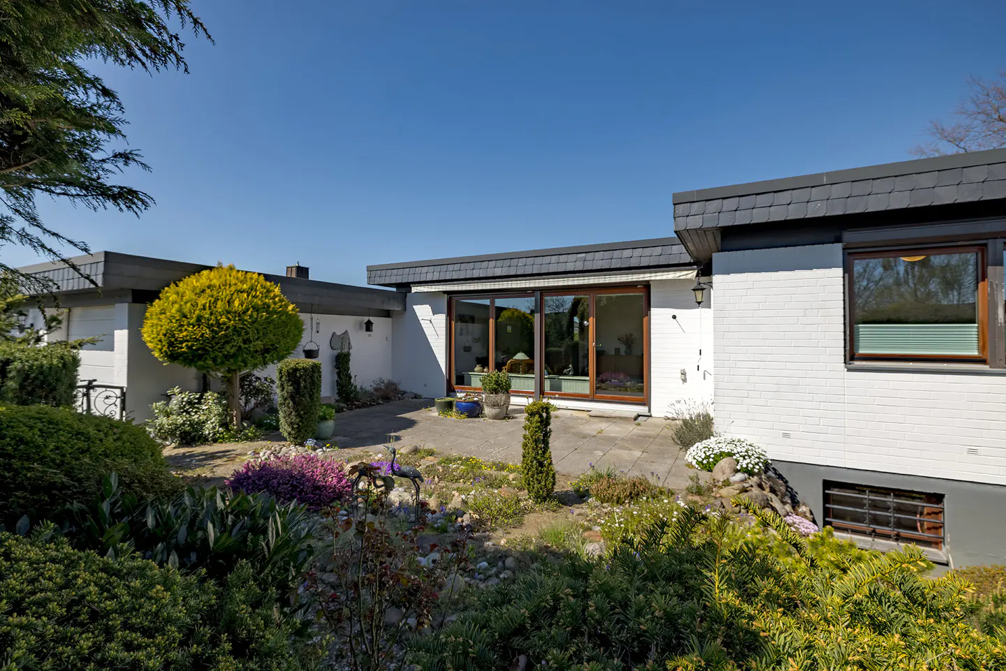 Exterior view of a white brick house with a gray roof and a stone patio surrounded by lush greenery.