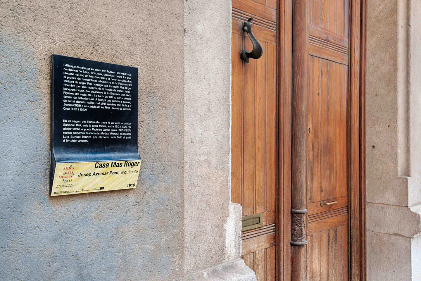 Close-up of a building's exterior with a wooden door, a metal knocker, and a plaque reading "Casa Mas Roger, Josep Azemar Pont, arquitecte, 1910."