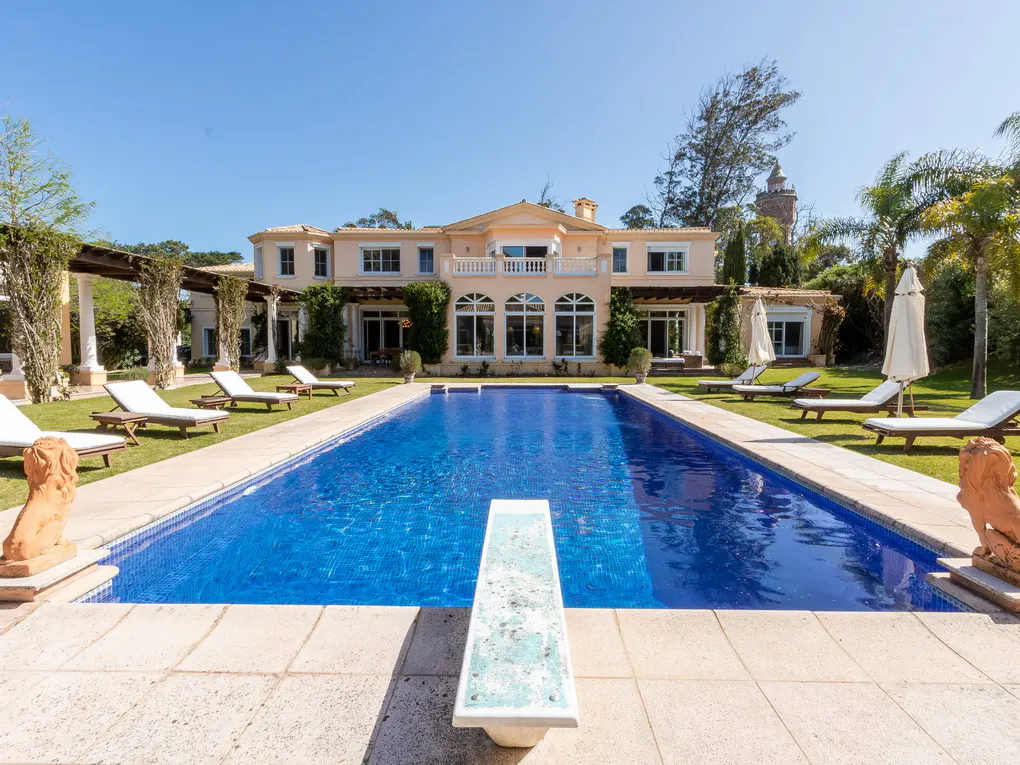 A large, blue, rectangular pool with a diving board in front of a tan two-story house. Lounge chairs and lion statues surround the pool.
