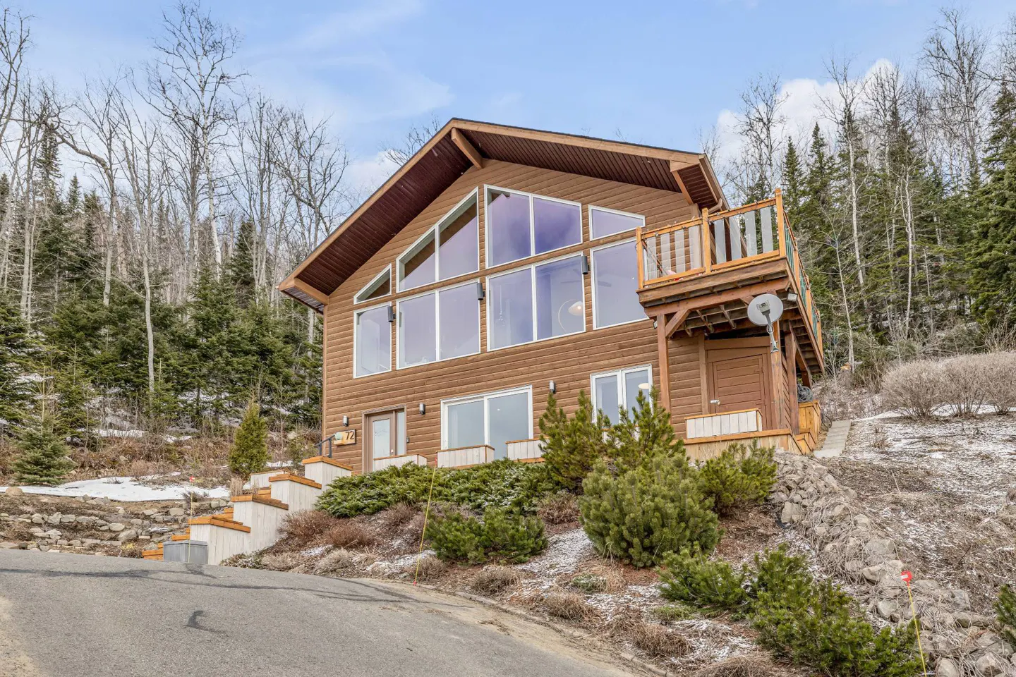 A brown, two-story house with a balcony sits on a hill surrounded by trees. The house has large windows and a brown roof.