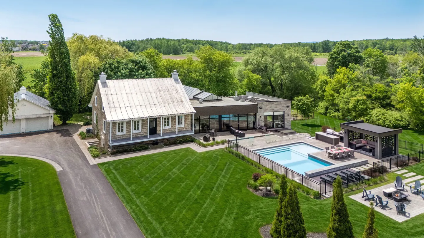 Aerial view of a stone farmhouse with a modern addition, pool, patio, and gazebo set in a lush, green rural landscape.
