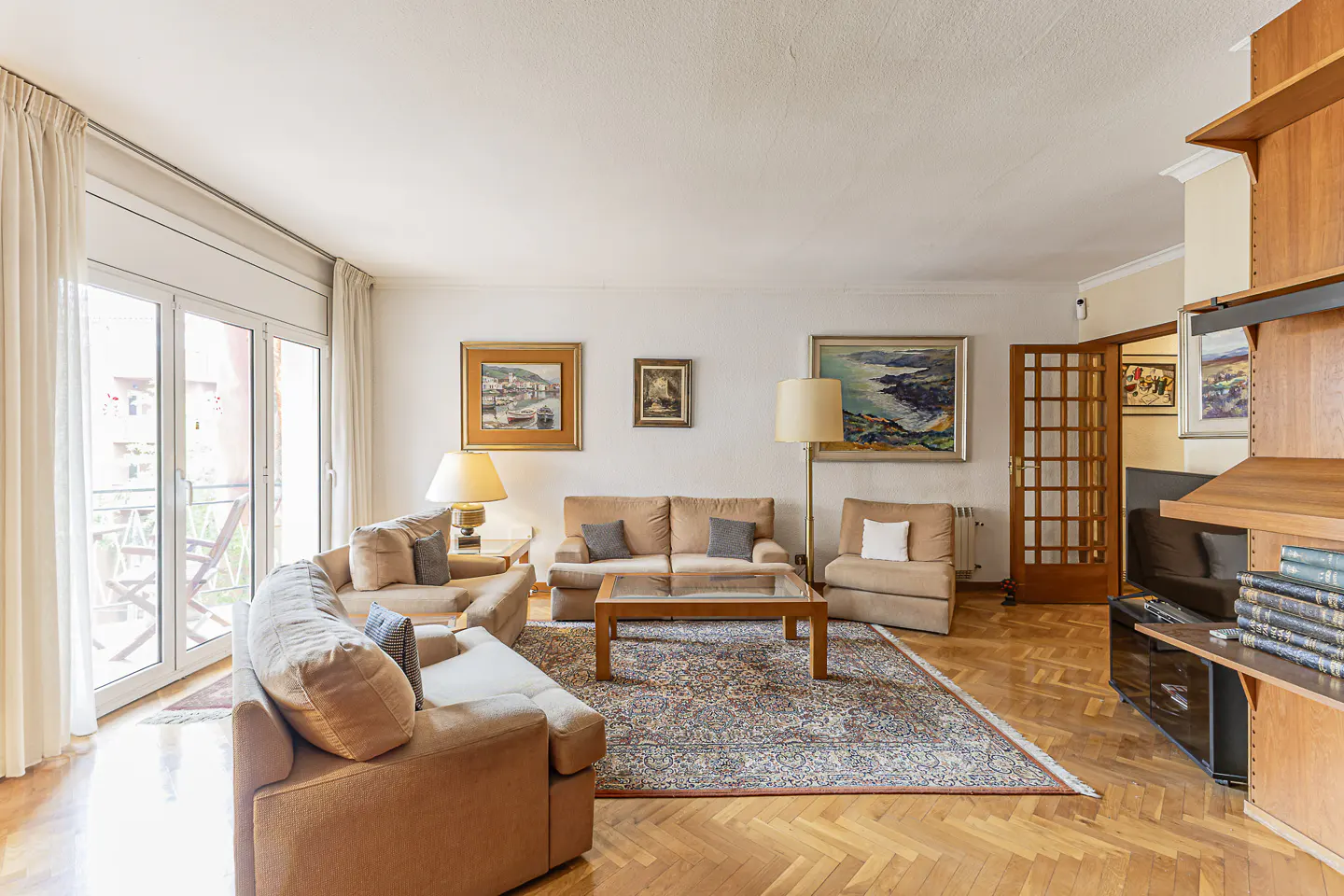 Bright living room with tan sofas, wood floors, and a patterned rug. Paintings adorn the walls, and a glass door leads to a balcony.