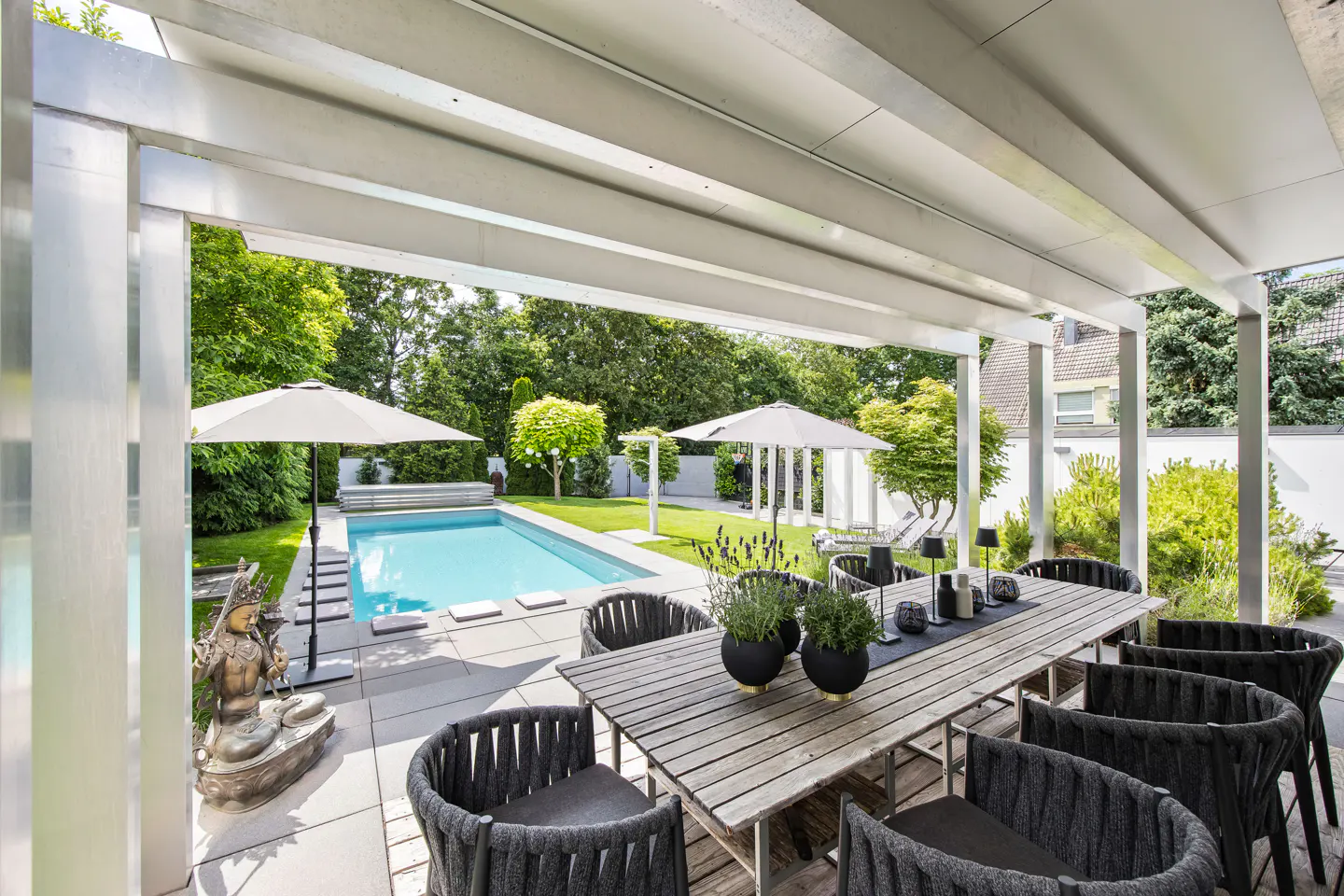 Covered patio with a wooden dining table and dark chairs overlooking a backyard swimming pool and lush green garden.