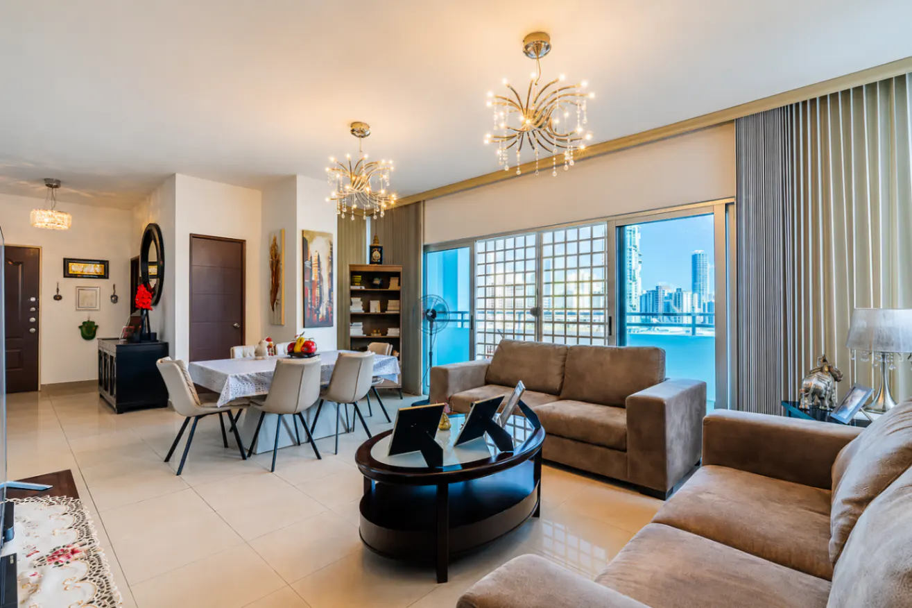 A modern living and dining area with brown sofas, a dining table, and a city view through a large window.