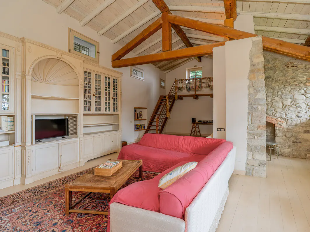 Bright living room with exposed beams, stone wall, and a large white bookcase. A red sofa and wooden coffee table sit on an ornate rug. Stairs lead to a loft.
