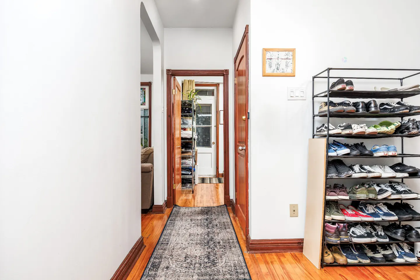 Hallway with hardwood floors, white walls, and a gray rug. A black metal shoe rack is filled with shoes. A doorway leads to another room.