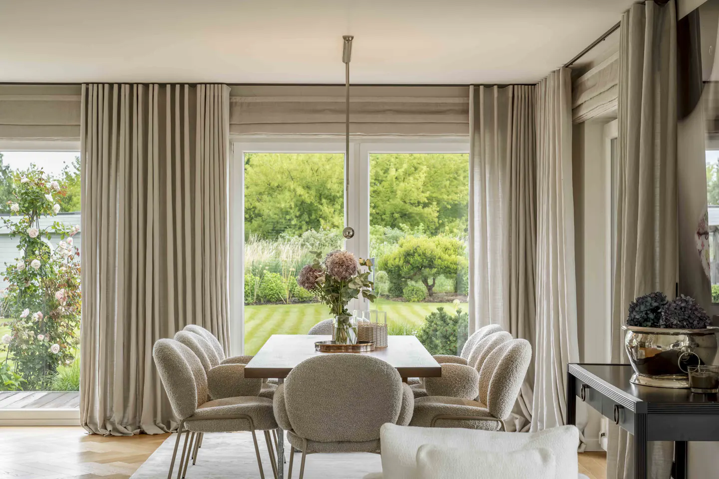 Dining room with a wood table, upholstered chairs, and beige curtains. A vase of flowers sits on the table, with a green garden view outside.