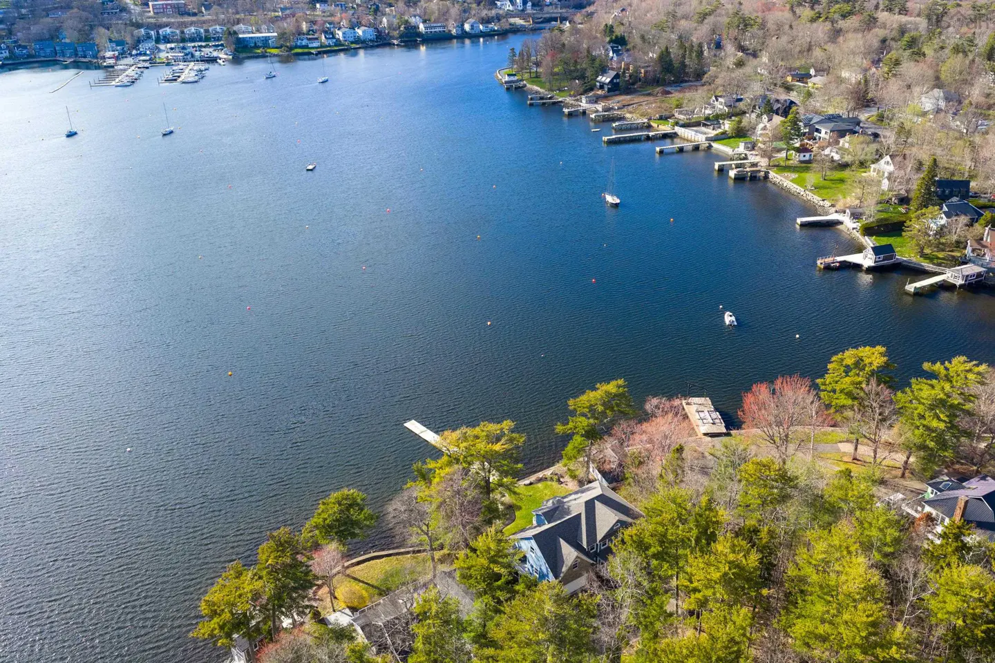 Aerial view of a blue house with a dock on a lake surrounded by green trees and other houses.