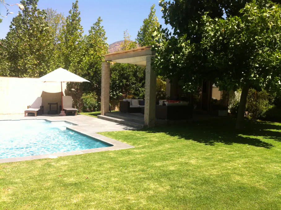 A backyard with a pool, lounge chairs, and a covered patio with outdoor seating. Green grass and trees surround the area.