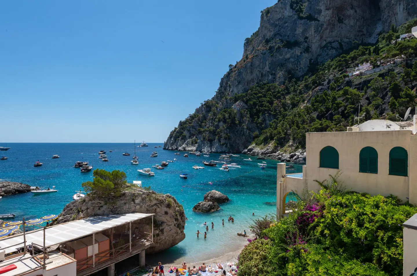 Coastal view of Capri, Italy, with turquoise water, boats, beachgoers, and a rocky cliffside under a clear blue sky.
