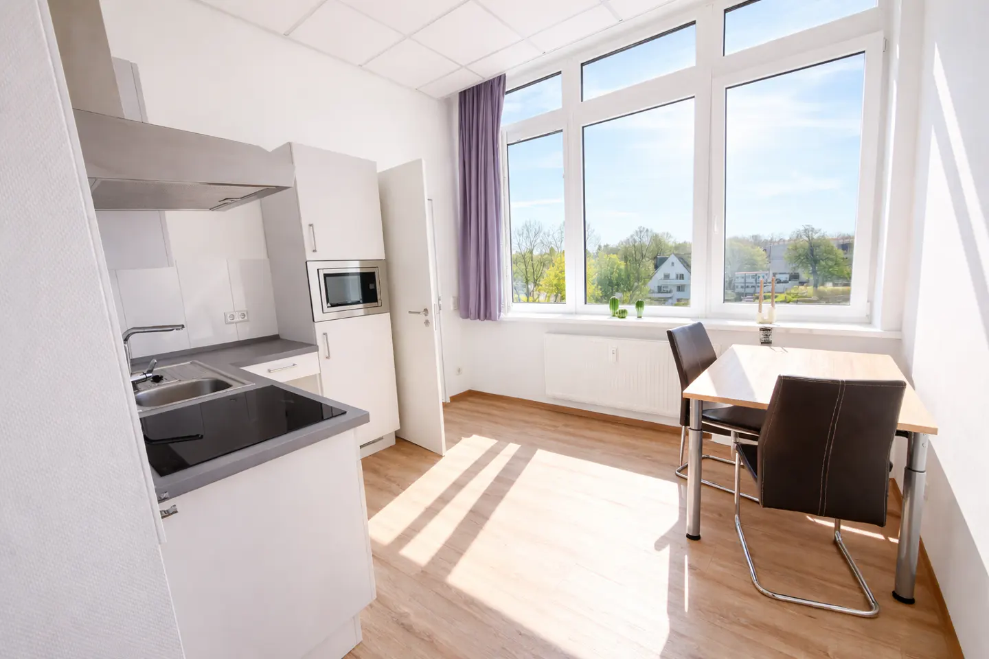 Bright studio apartment with white kitchen cabinets, a dining table with two chairs, and a large window with a view of trees.