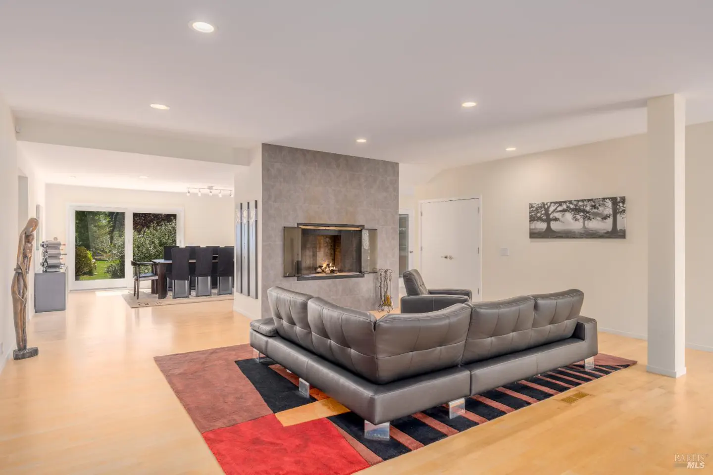 Bright living room with a gray leather sofa, fireplace, and a colorful geometric rug on a light wood floor. Dining room visible in the background.