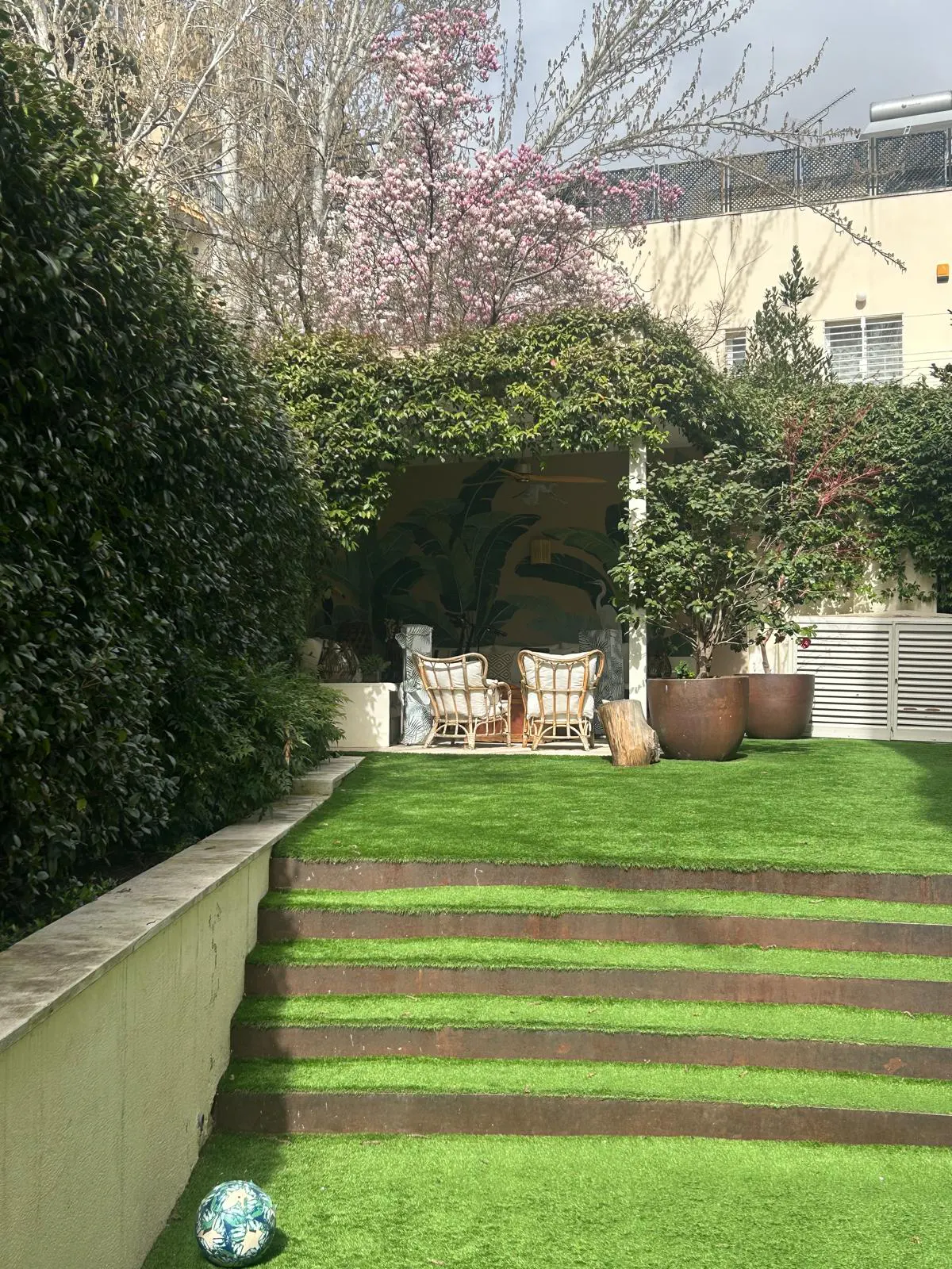 A lush backyard with tiered grass steps, a covered patio with two wicker chairs, and a blooming pink tree in the background.