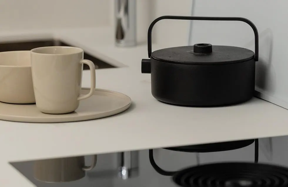 Close-up of a modern kitchen counter with a black pot, beige mug, and bowl on a plate. The counter is white, and a black stovetop is visible.