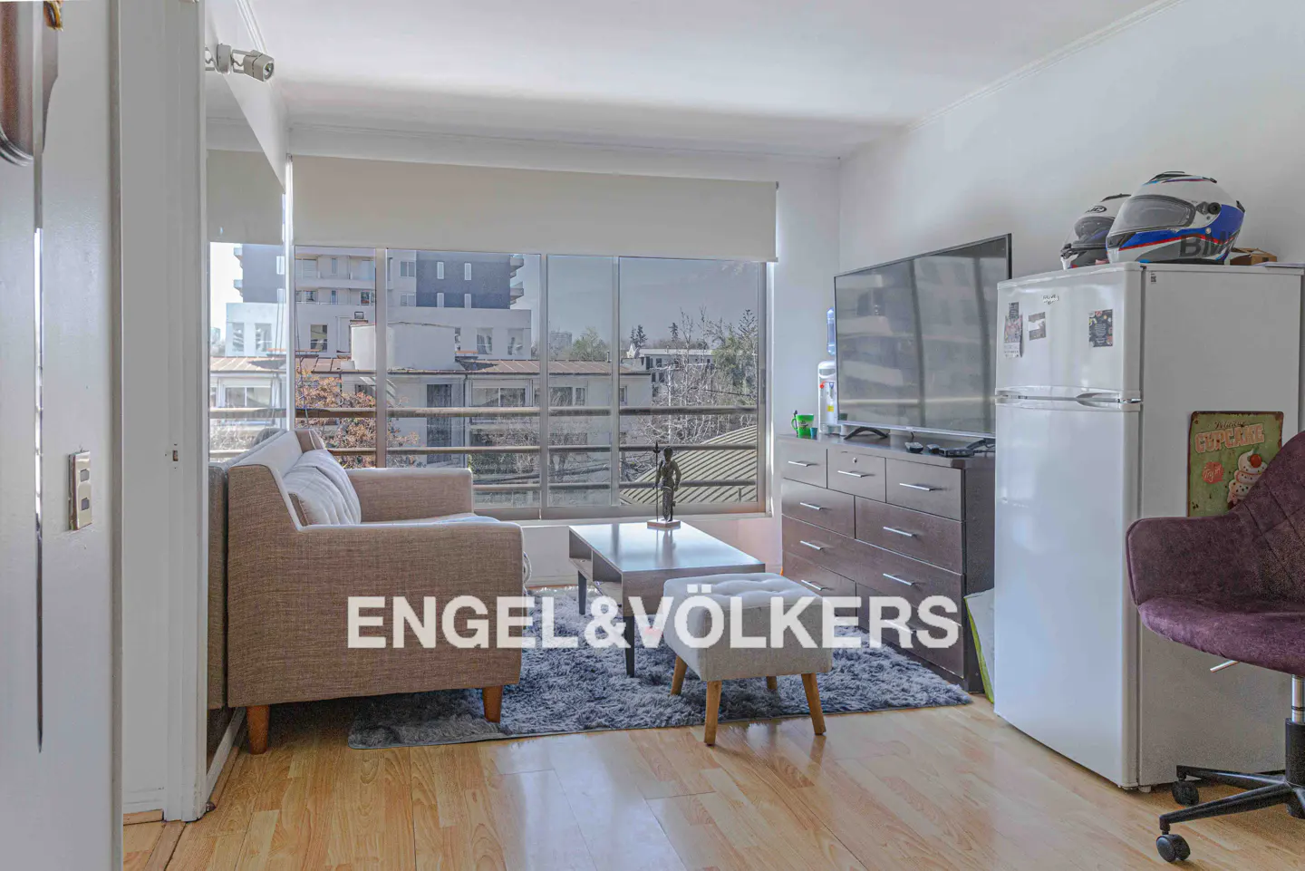 Living room with a beige sofa, coffee table, TV, dresser, white fridge, and purple chair. Large windows overlook city buildings.